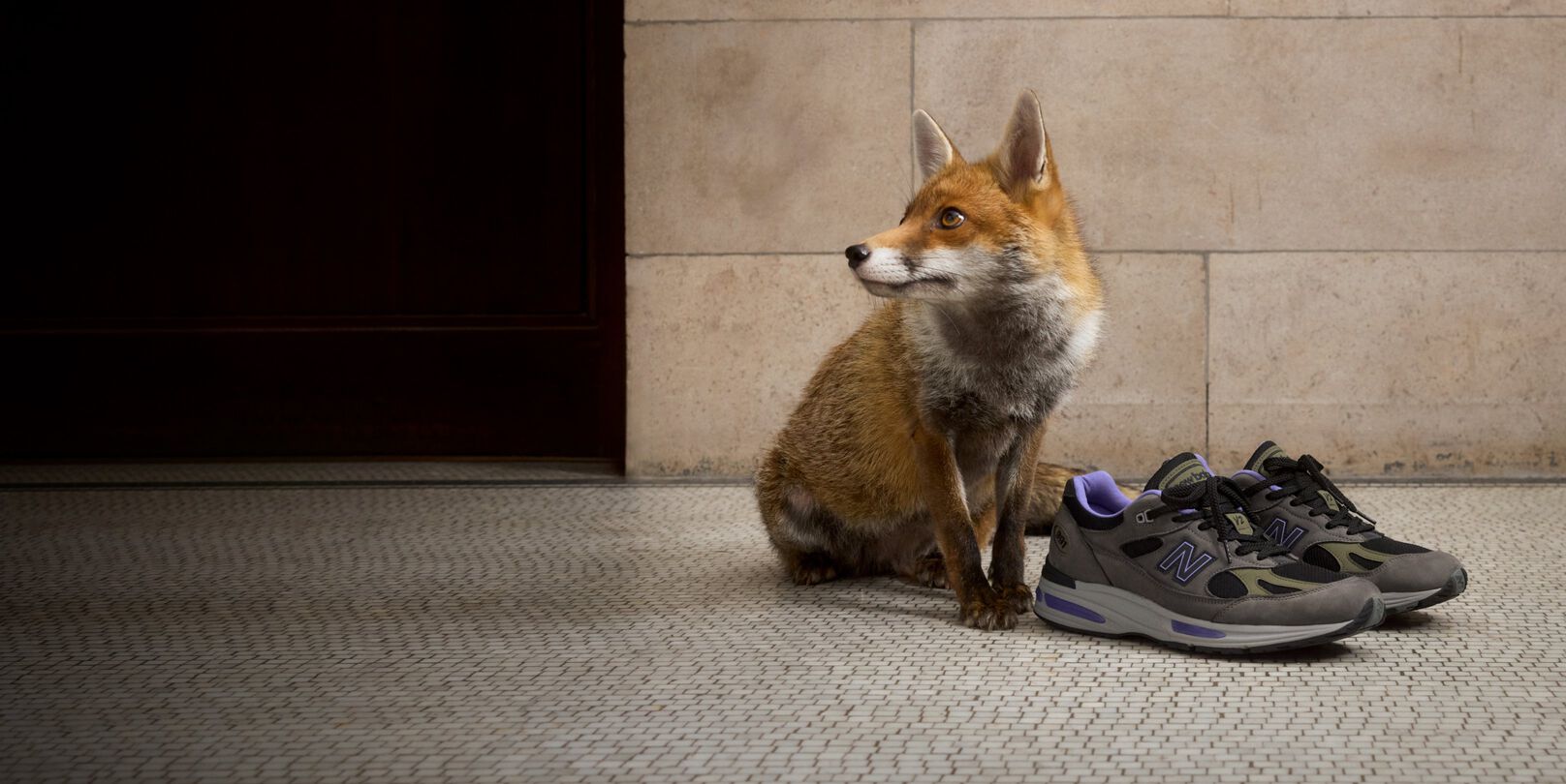 A pair of New Balance Made in UK sneakers on a white tiled floor with stone walls in the background next to a sitting fox. 