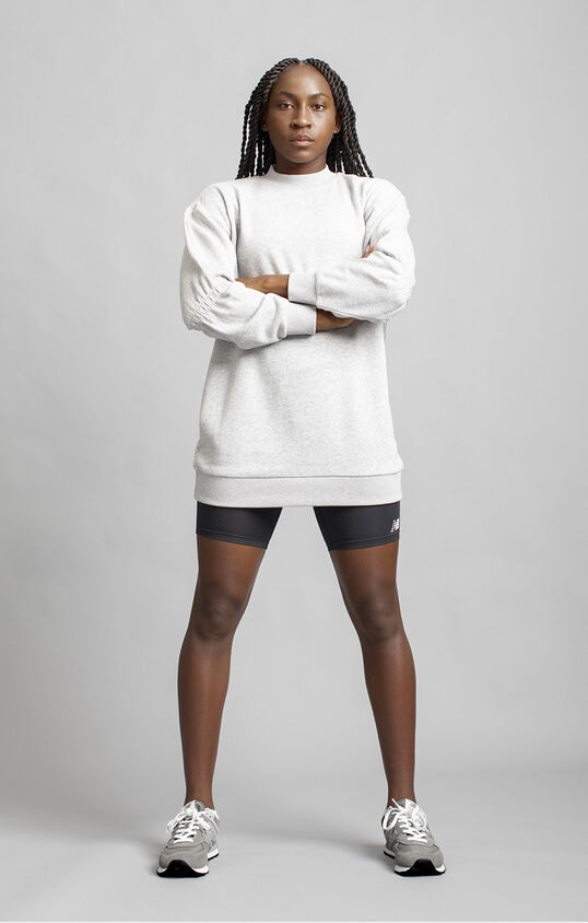Coco Gauff standing against grey studio backdrop with arms crossed while wearing grey NB apparel and shoes.