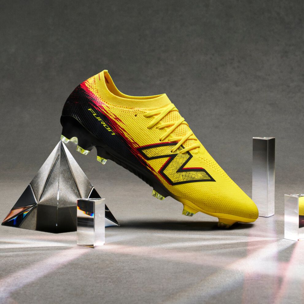 A bright yellow soccer cleat with black and red accents is displayed on its side among several glass prisms on a neutral gray surface.