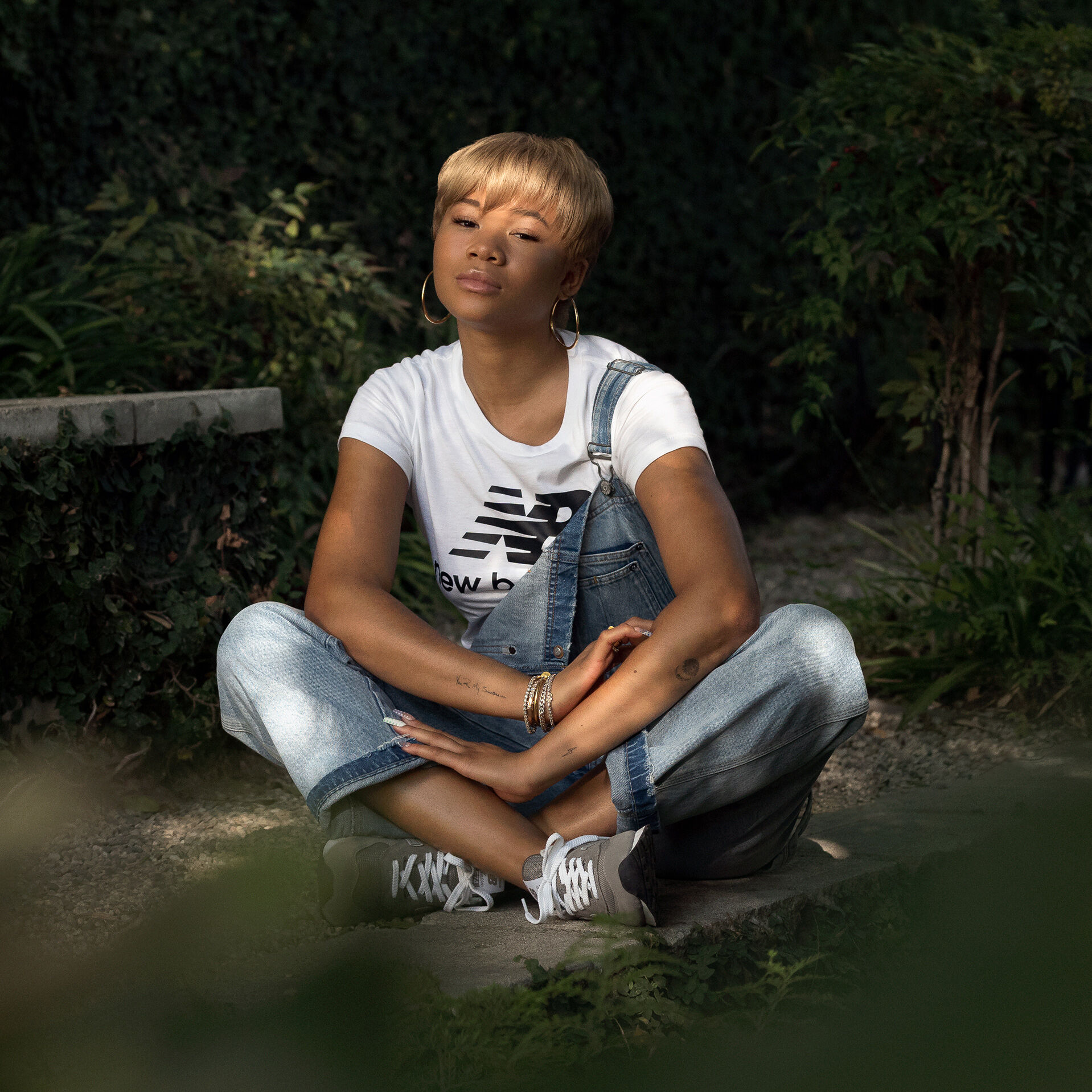 Storm Reid wearing a white New Balance logo tee and light blue overalls, sitting on the ground surrounded by greenery
