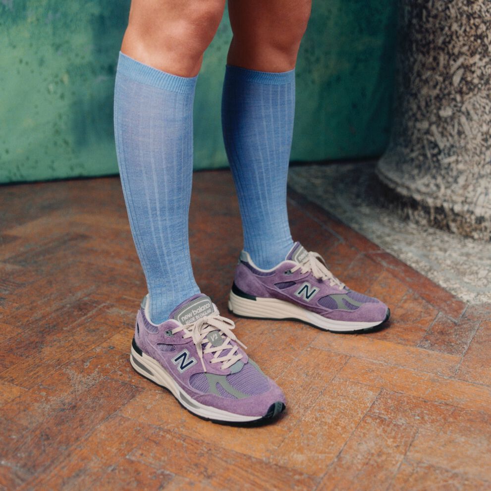 A pair of New Balance Made in UK sneakers standing on a wooden floor with textured green walls and a stone pillar in the background. 