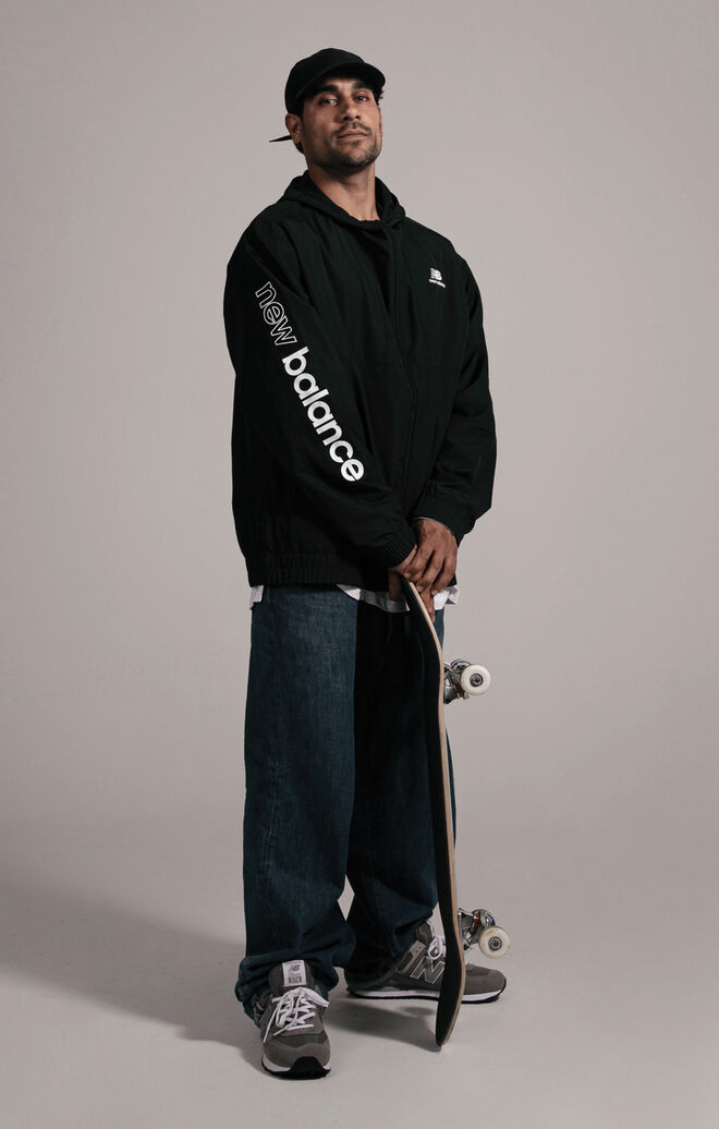 Tiago Lemos standing against grey studio backdrop holding his skateboard while wearing grey NB apparel and shoes 