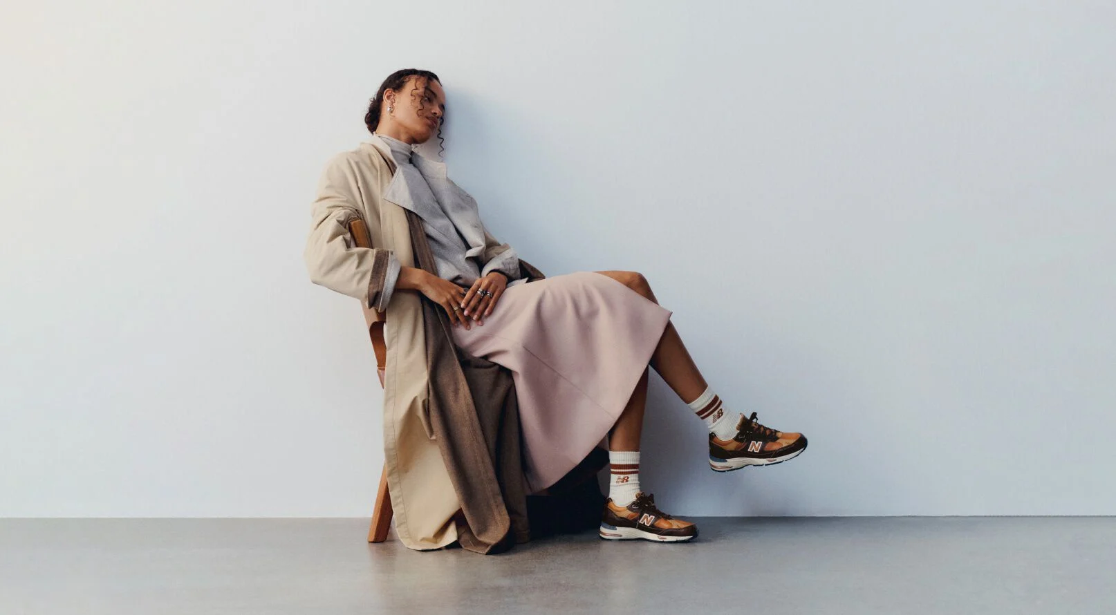A woman sitting on a wooden chair with her legs crossed wearing a pair of New Balance Made in UK sneakers in a minimalist room with a white wall and concrete floor